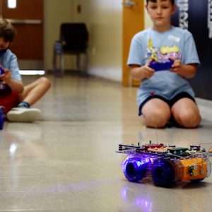 Two boys at robotics summer camp sitting on the floor with remote controls piloting their robots.