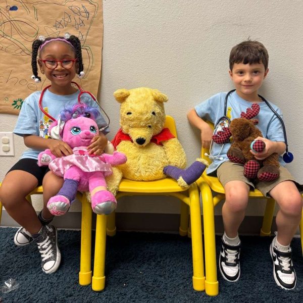 Two smiling children in blue shirts sit on yellow chairs in a clinic, each with stuffed toys (pink bunny and Pooh bear).